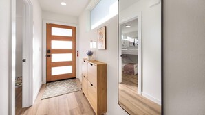 Ein Flur mit einer Holztür, einem Spiegel, der ein Zimmer mit einem Bett reflektiert, und einer kleinen Holzkommode mit einer Vase.