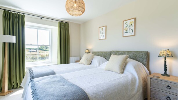 Ein Doppelbett mit weißblauer Decke, zwei Nachttischlampen, ein Nachttisch und ein Fenster mit grünen Vorhängen.