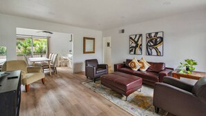 Amplia sala de estar con piso de madera, sofá de cuero marrón, zona de comedor con mesa y sillas blancas, y dos cuadros abstractos en la pared.