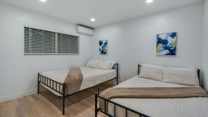 Una habitación con dos camas, una ventana con persianas, un equipo de aire acondicionado y dos cuadros abstractos en las paredes.