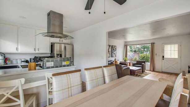 Una cocina moderna con armarios blancos, un refrigerador de acero inoxidable y un horno empotrado. Hay un comedor con una mesa grande y sillas, y un ventilador de techo.