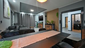 A modern living room with a large TV, a dining table with a pink runner, and a kitchen area visible through an open door.