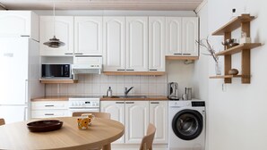 Une cuisine moderne avec des meubles blancs, un four à micro-ondes, une cuisinière et un lave-linge. On y trouve une table à manger en bois, des chaises et un saladier.