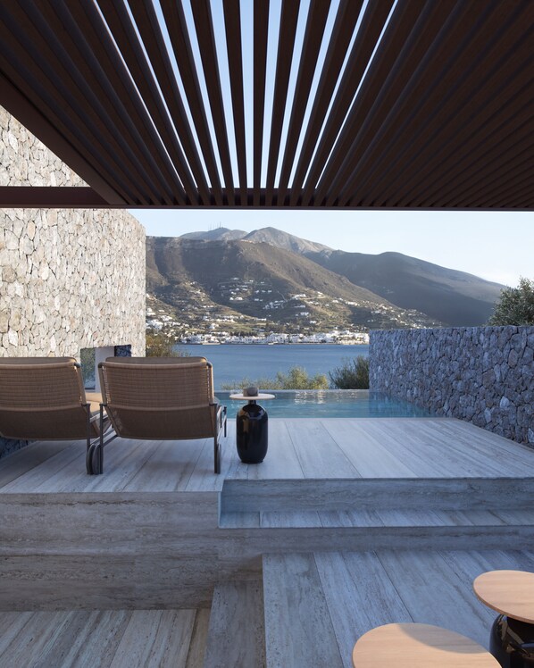 Un espace extérieur moderne, aménagé avec un mur en pierre, des lambris en bois et offrant une vue sur un paysage montagneux et une étendue d’eau.