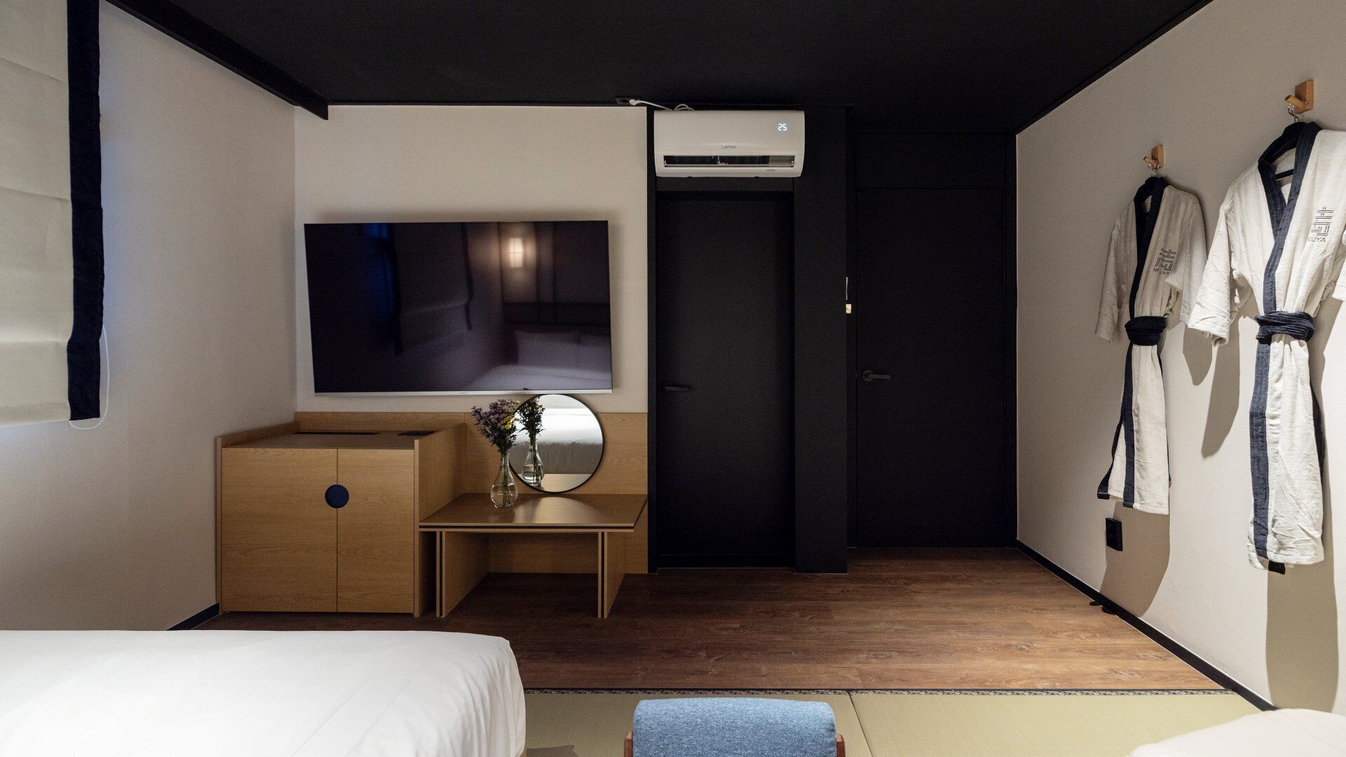 A modern hotel room with a large flat-screen TV, a wooden cabinet, a small table with a vase, and a pair of white robes hanging on the wall.