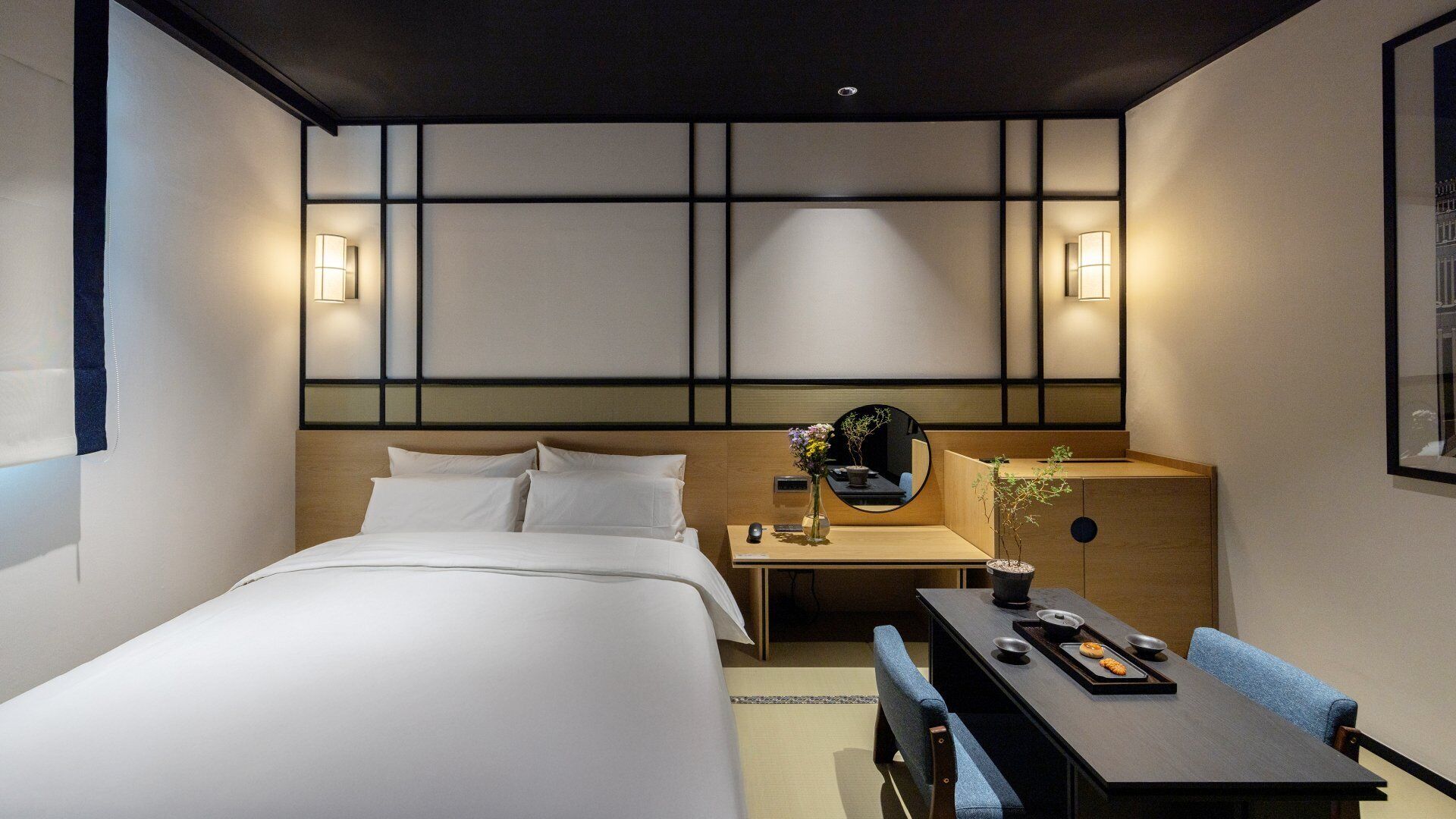 A modern hotel room with a neatly made bed, a bedside table with a lamp, a small desk with a chair, and a wall-mounted mirror.