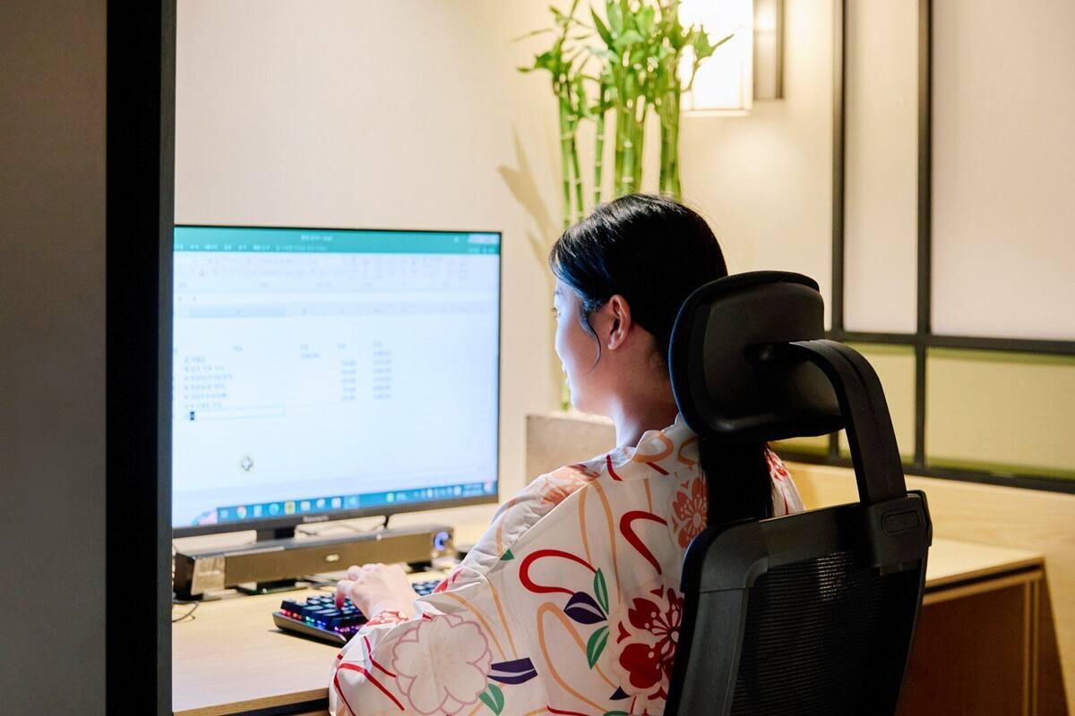 A person seated at a desk with a computer, a keyboard, and a mouse. There is a plant in the background.
