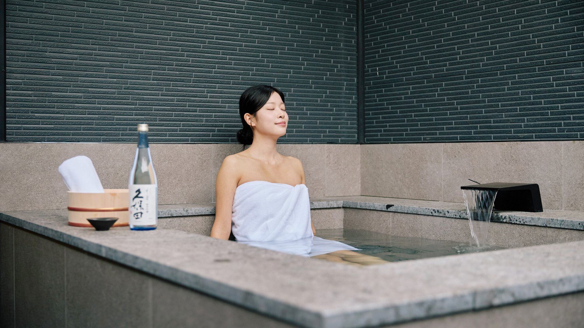 Una persona en un baño de spa con una botella de sake y una toalla.