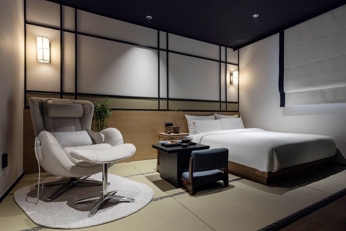 A modern bedroom with a bed, a chair, a small table, and wall-mounted lighting.