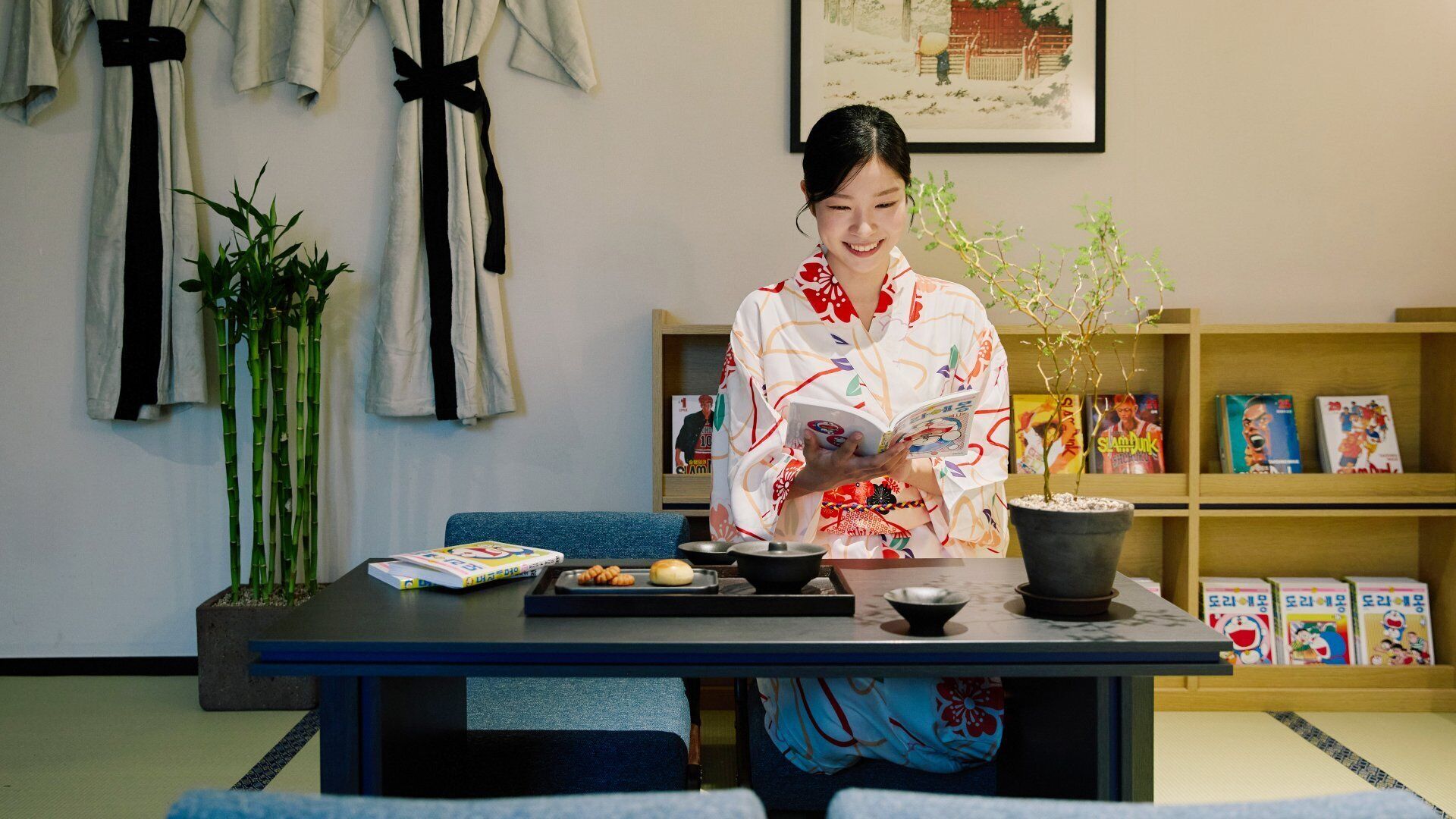 Ett rum med en traditionell japansk inredning, med tatamimattor på golvet, ett lågt bord med ett teservis och en bokhylla fylld med böcker och tidskrifter.