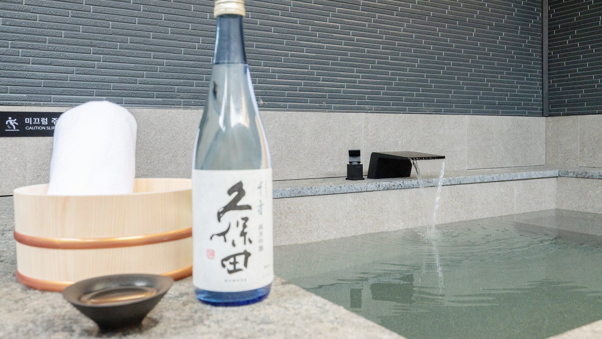 Eine Sake-Flasche, ein Holzsake-Set und eine Steinschale auf einer Steinplatte neben einem Wasserspiel.