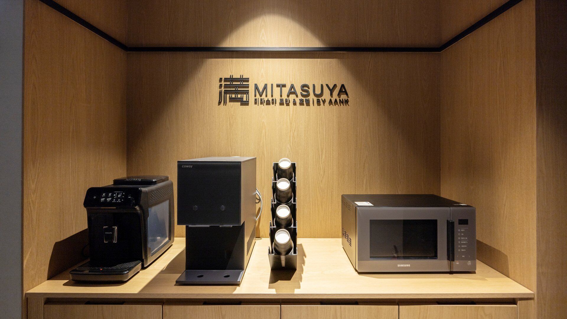 A display of electronic appliances including a coffee maker, a speaker, and a microwave, with a wooden wall and a sign reading "MITASUYA".