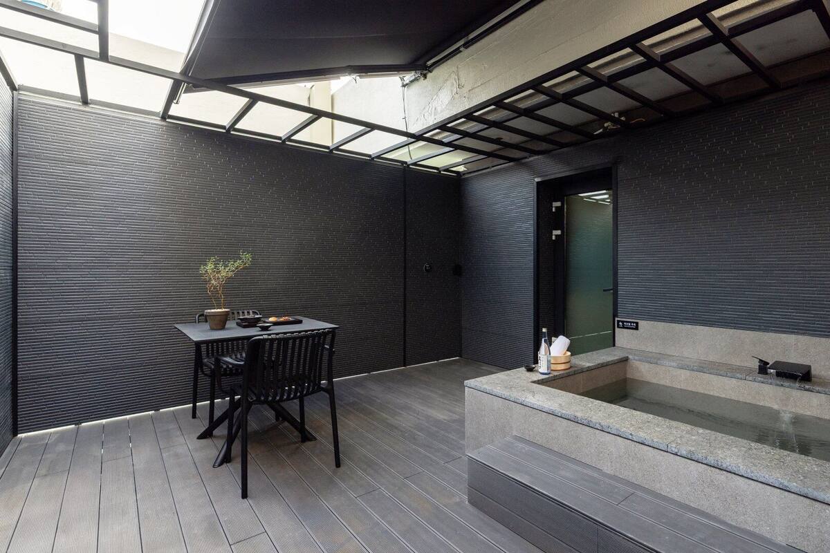 A modern outdoor patio with a dining table and chairs, a small plant, and a stone basin with a water feature.