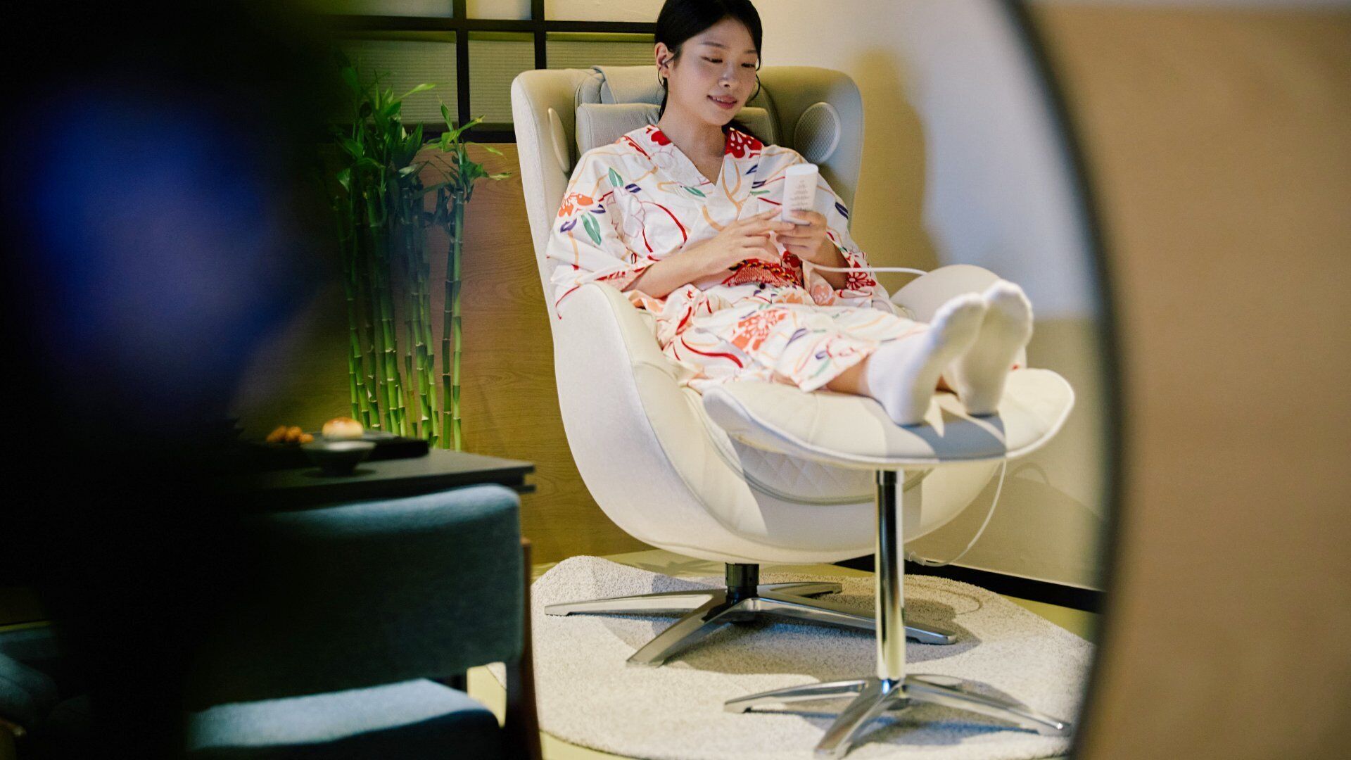 Une personne assise sur une chaise blanche dotée d’un repose-pieds, vêtue d’une robe à motifs floraux, se trouve dans une pièce où se trouvent un plant de bambou et une petite table sur laquelle est posé un bol.