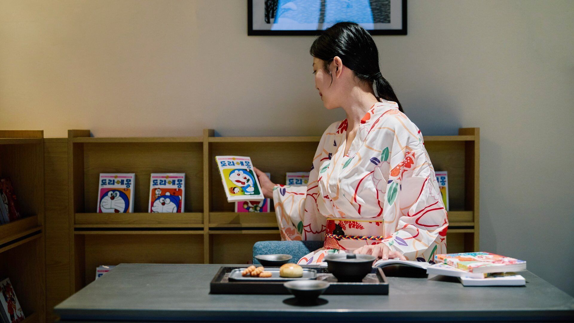 A person seated at a table with a book, a bowl, and some food items. There are shelves with books and a framed picture on the wall.