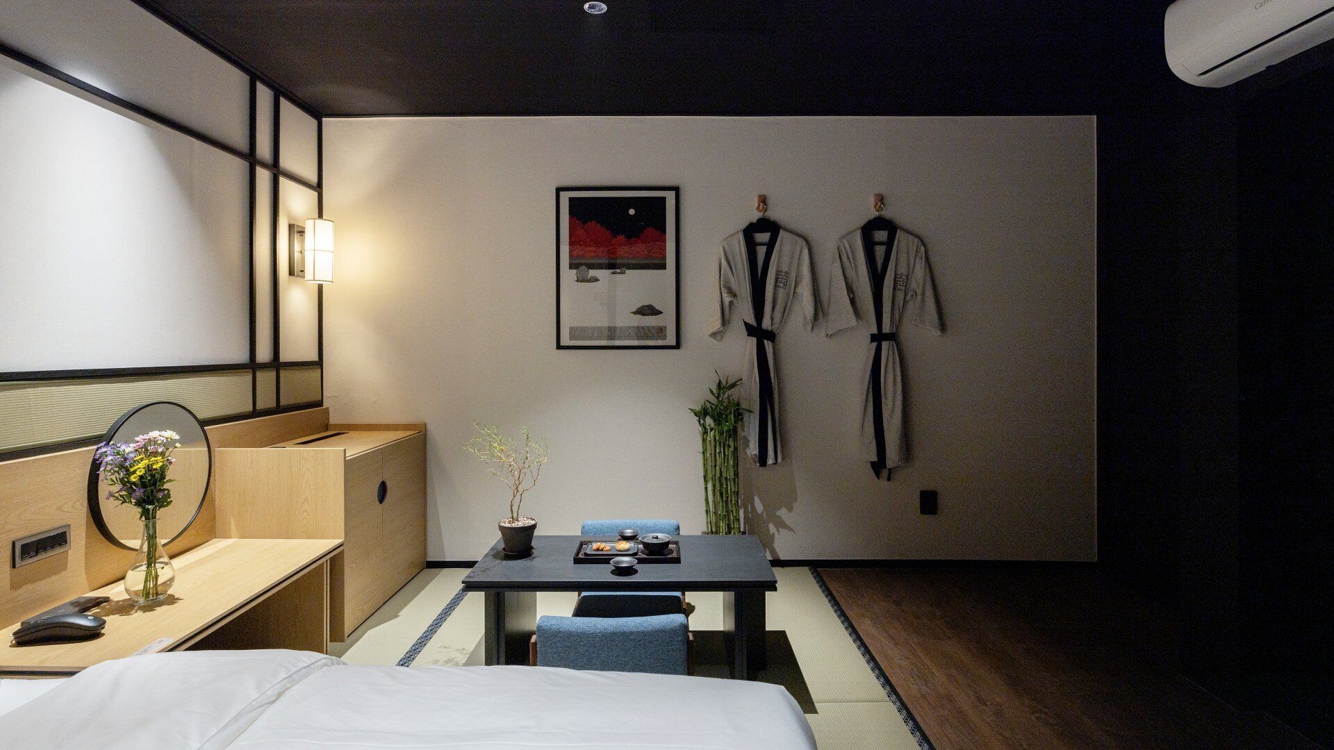 Ein Hotelzimmer mit Bett, Nachttisch, Spiegel und kleinem Schreibtisch mit Stuhl. An der Wand hängt ein gerahmtes Bild und zwei Bademäntel hängen an Haken.