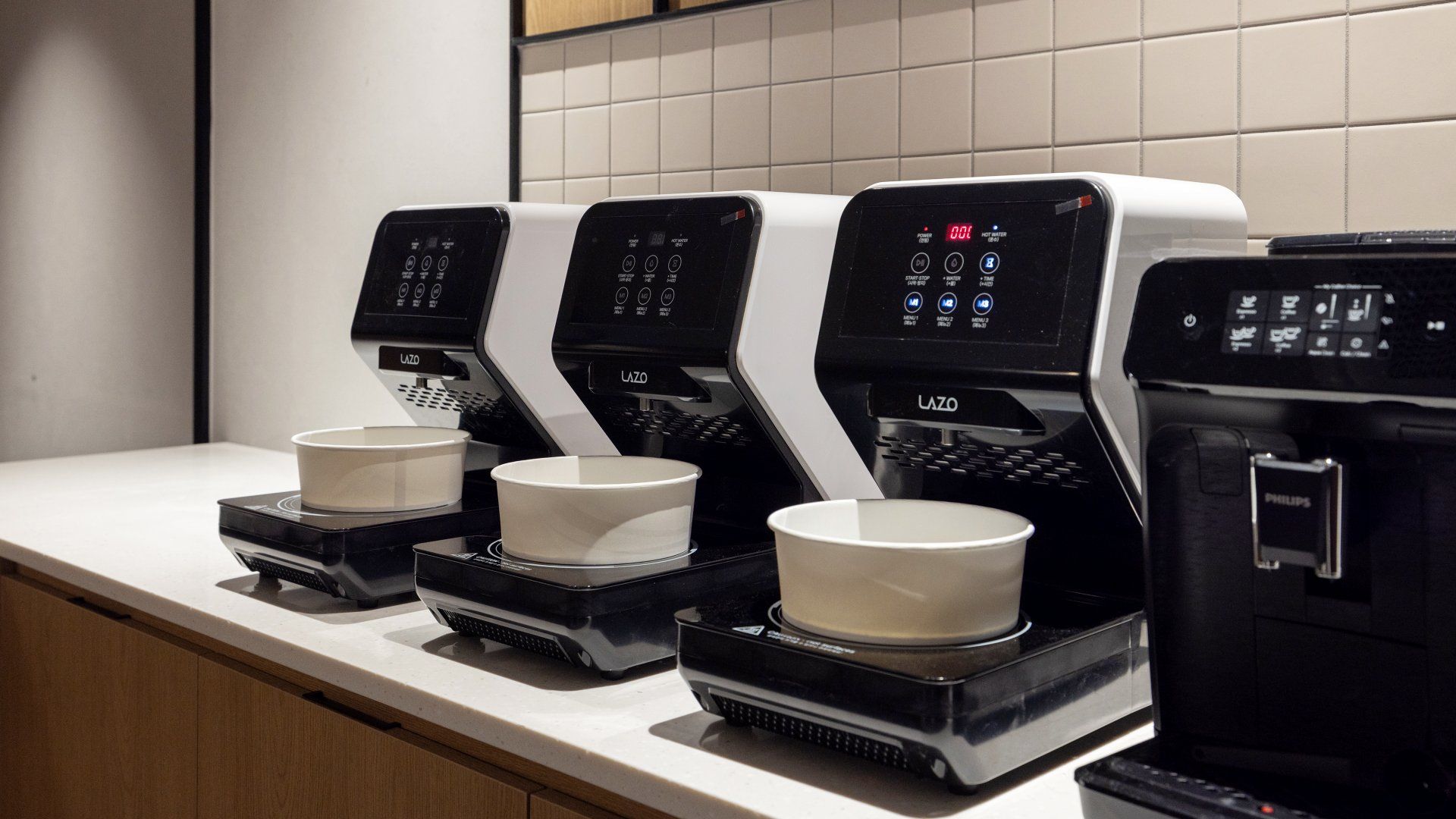 Tres máquinas de café en fila sobre una encimera de cocina, con tazas colocadas en sus bandejas.