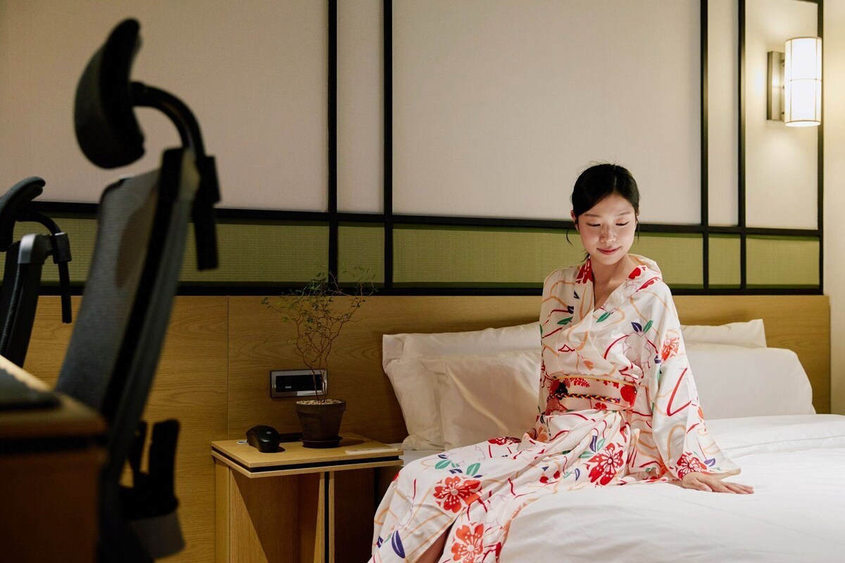 A hotel room with a bed, a bedside table with a plant, a lamp, and a chair.
