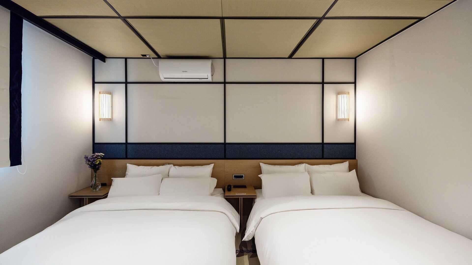Ein Hotelzimmer mit zwei Betten, einem Nachttisch, einer Vase mit Blumen und einer Klimaanlage in der Decke.