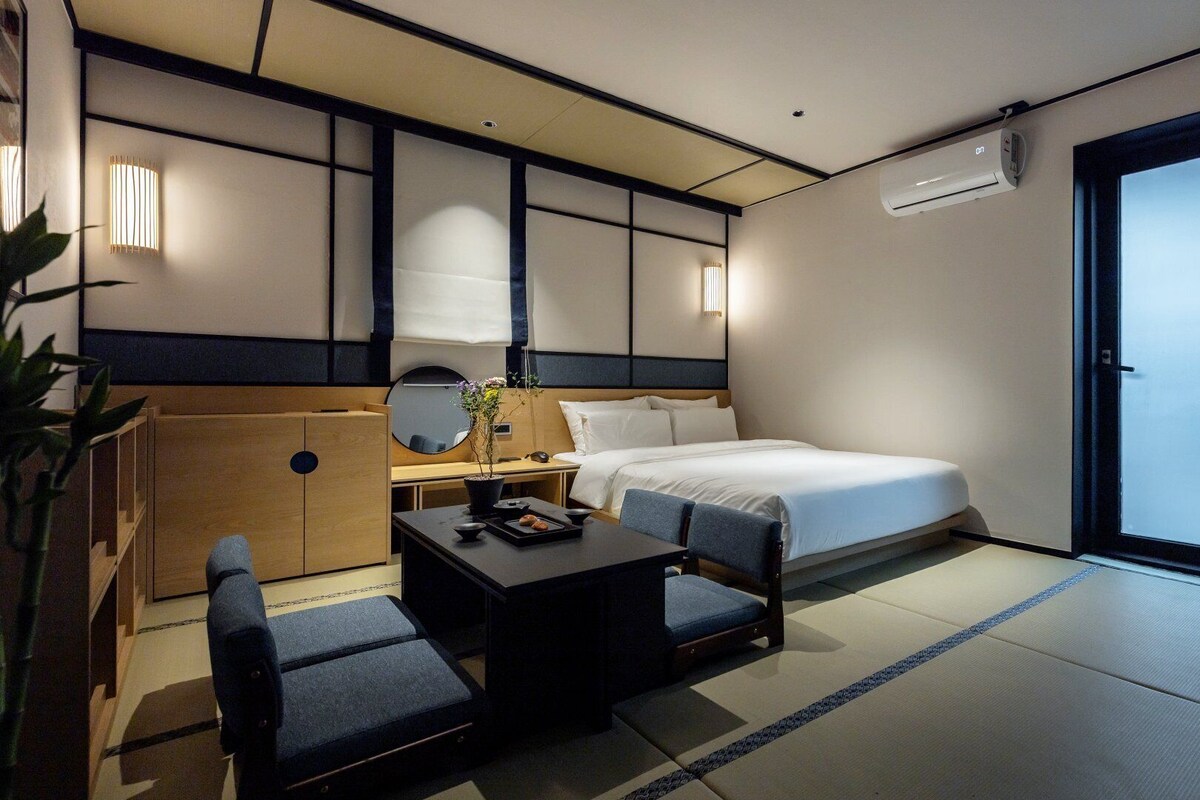A hotel room with a bed, a desk, a chair, a small table, a mirror, and a potted plant.
