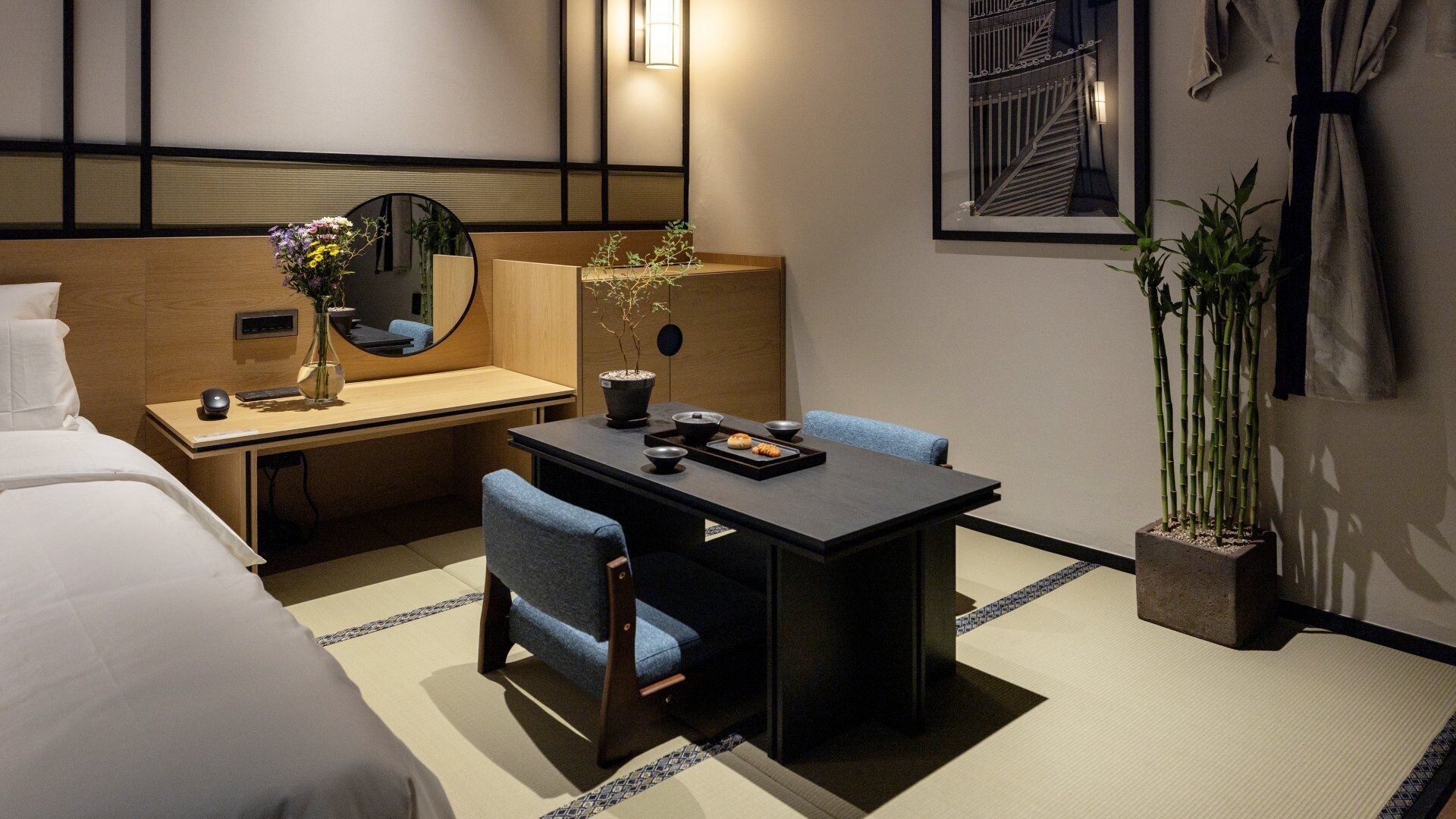 A hotel room with a bed, a desk with a mirror, a small table with chairs, a potted plant, and a framed picture on the wall.