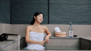 A person sitting on steps with a towel wrapped around their body, a wooden box with Japanese characters, and a bottle on a ledge. - Mitasuya Ryokan Hotel by Anook Pyeongtaek Station (Pyeongtaek)