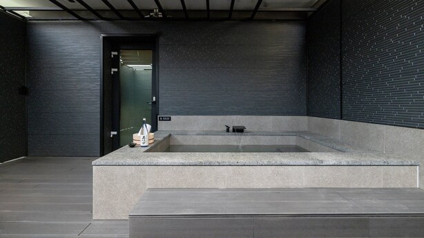 A modern bathroom with a rectangular bathtub, a small sink, and a toilet. There is a door leading to another room, and the walls are covered with dark tiles. - Mitasuya Ryokan Hotel by Anook Pyeongtaek Station (Pyeongtaek)
