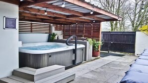 Un jacuzzi bajo una pérgola de madera, con escalones que llevan a él, rodeado de un jardín cercado.