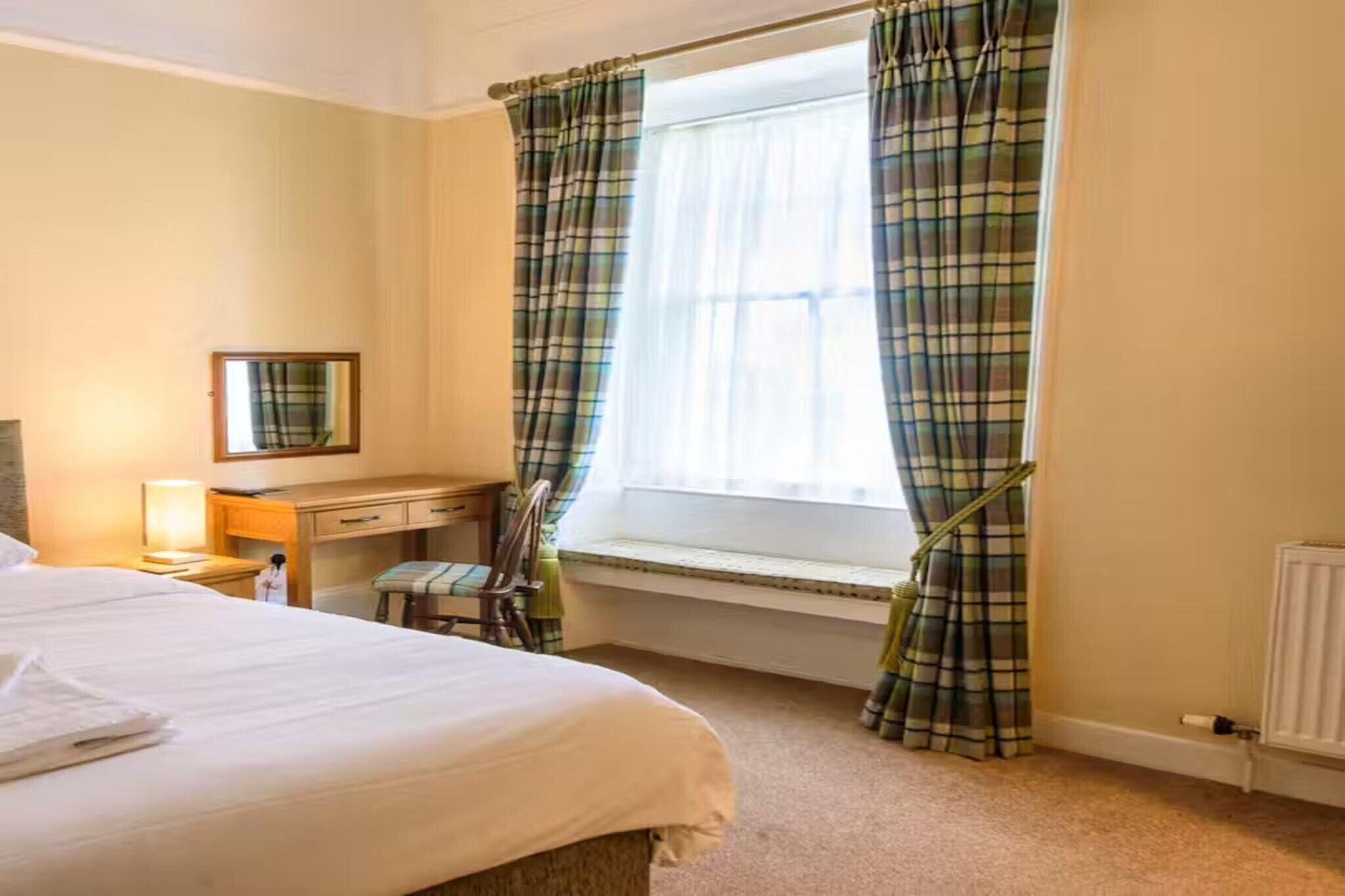 Ein Hotelzimmer mit Bett, Schreibtisch, Stuhl, Fenster mit Karottenvorhängen und Heizkörper.