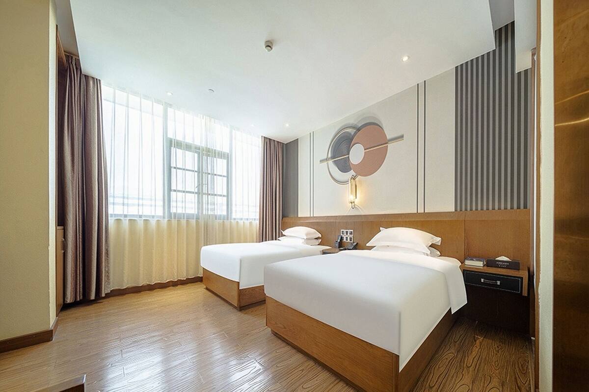 A hotel room with two beds, a large window with curtains, a wall-mounted clock, and a bedside table with a phone and a book.