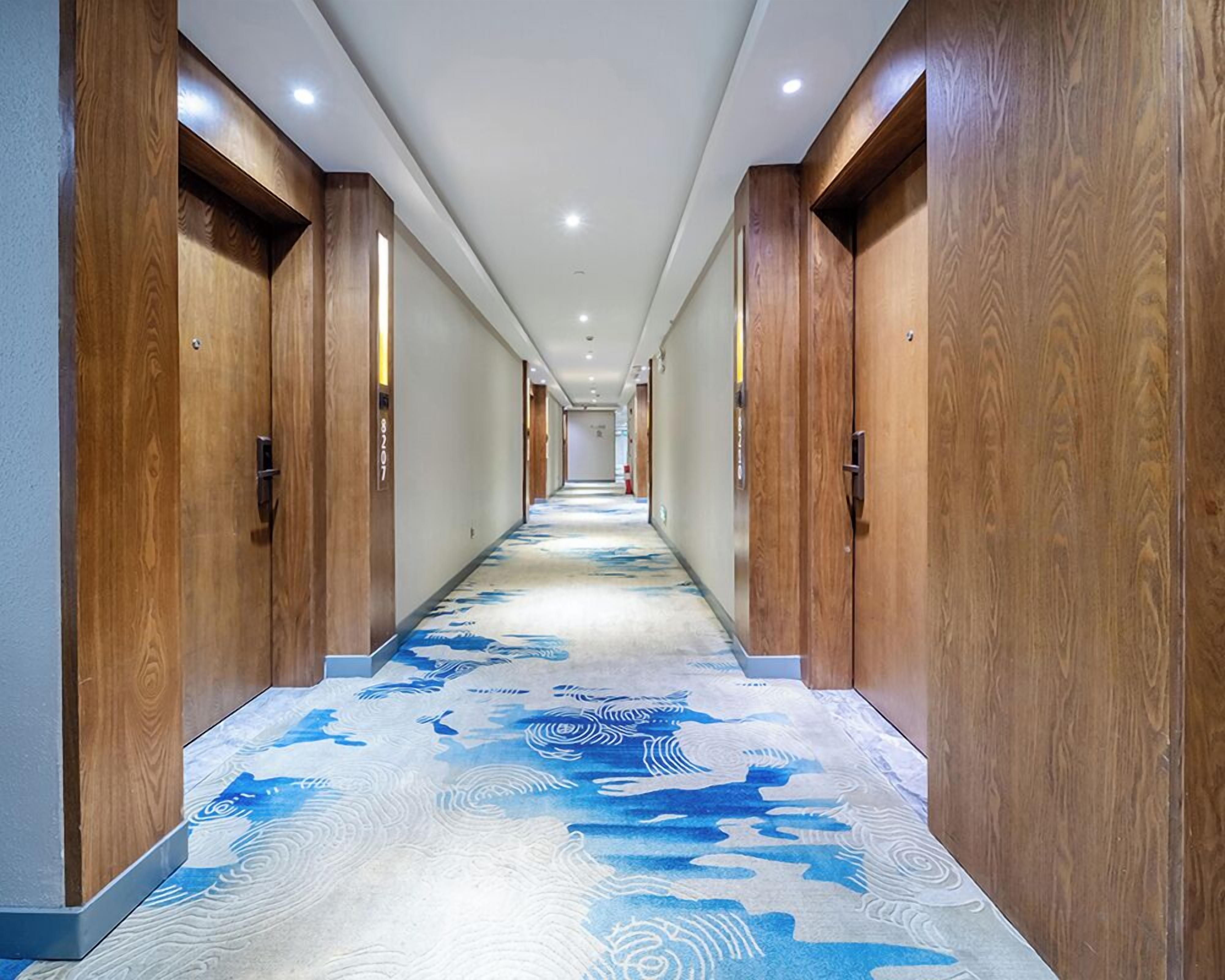 A hotel hallway with wooden doors, a patterned carpet, and a ceiling with recessed lighting.