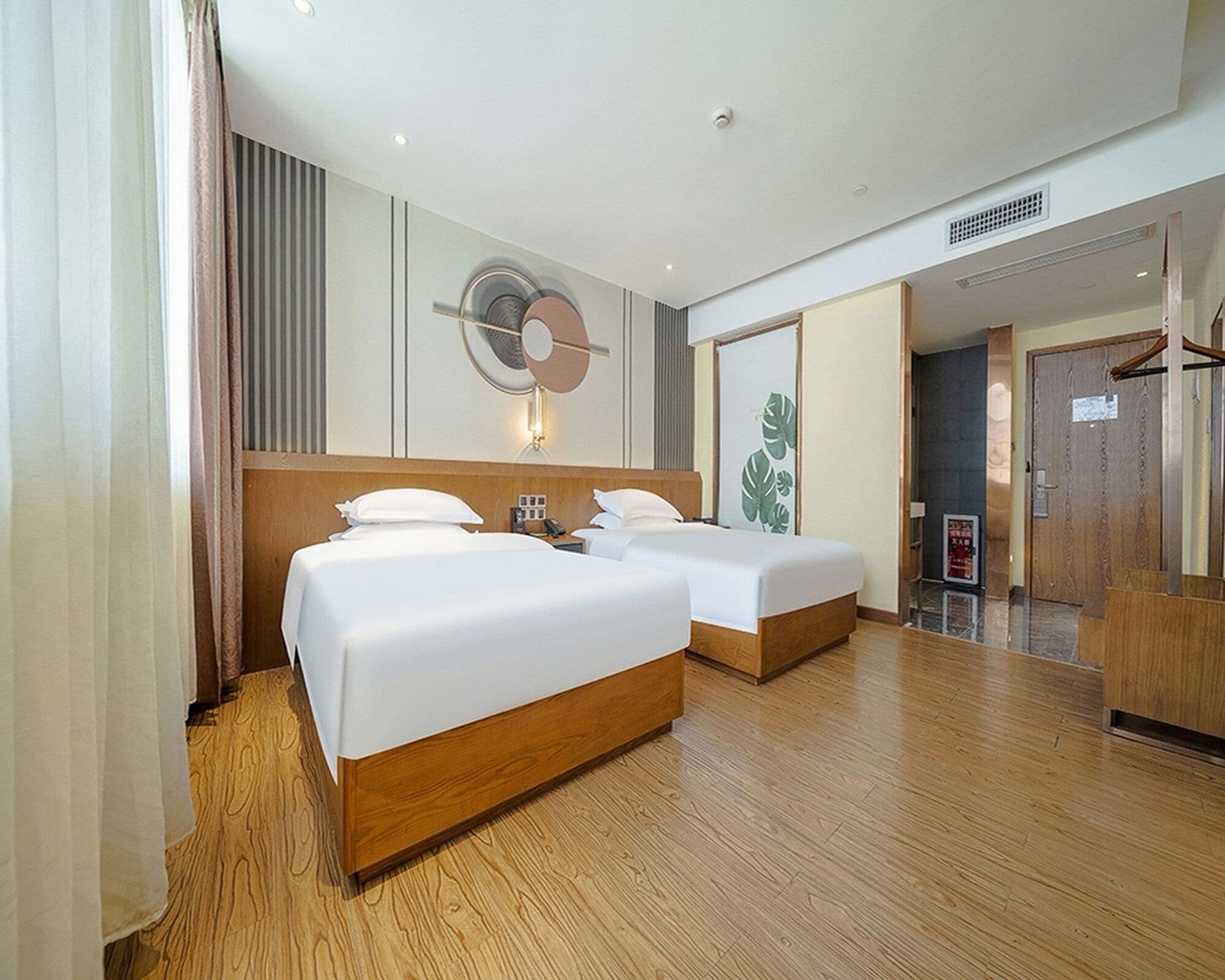 A hotel room with two beds, a wooden floor, a wall-mounted fan, and a door leading to a hallway.