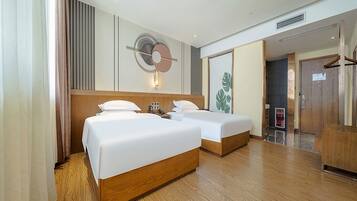 A hotel room with two beds, a wooden floor, a wall-mounted fan, and a door leading to a hallway.