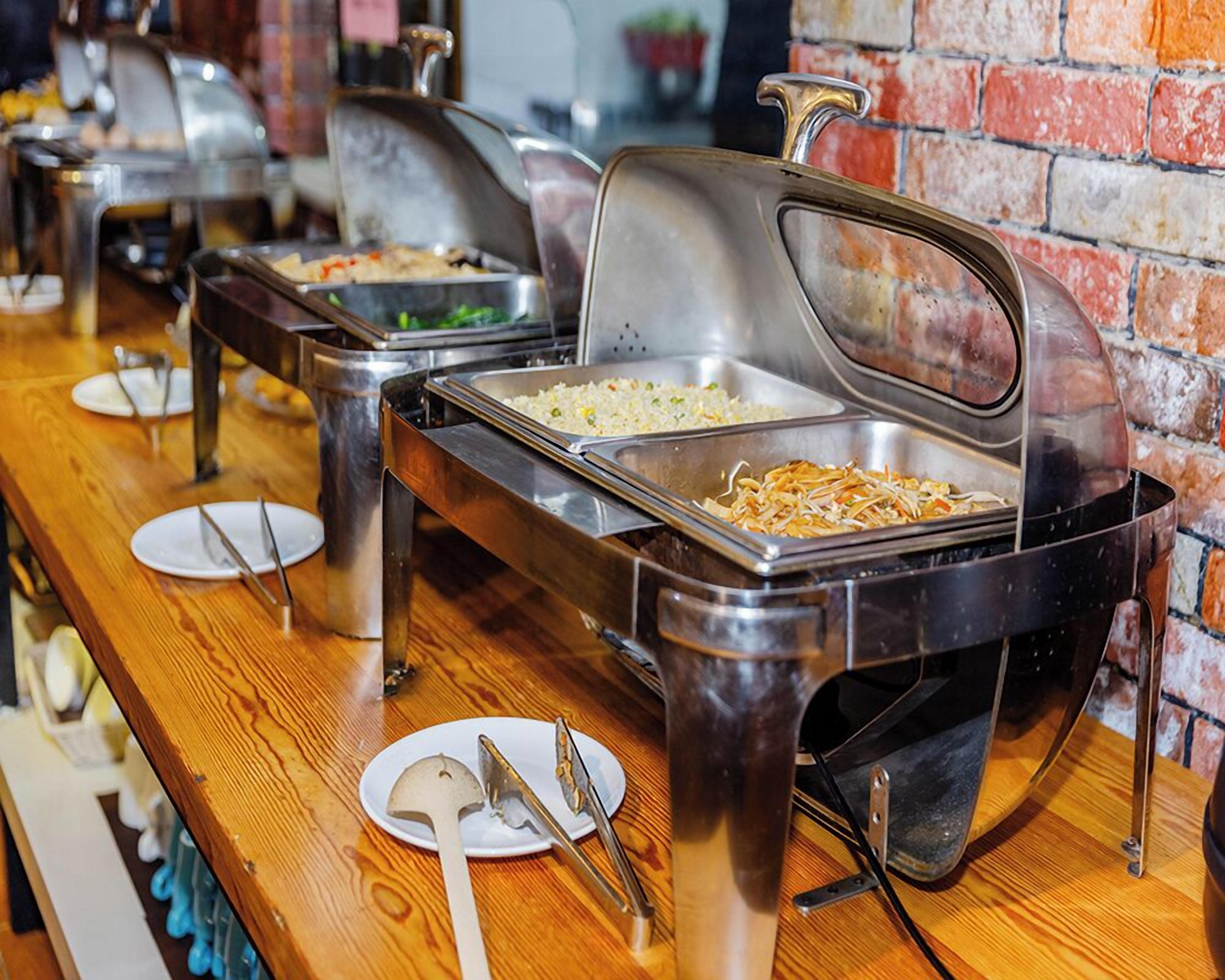Ein reichhaltiges Buffet mit verschiedenen Speisen in Metallservierplatten, auf einem Holztisch vor einer Ziegelwand präsentiert.