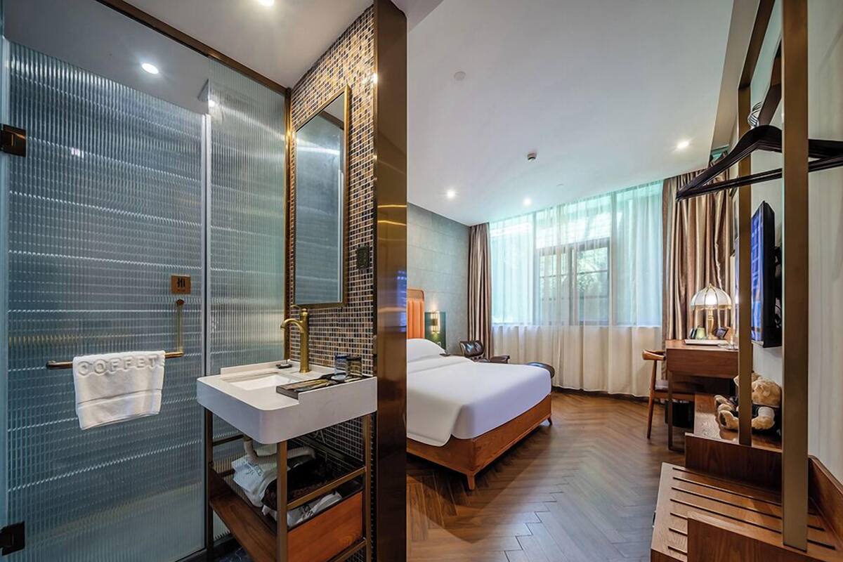 A modern hotel room with a glass shower enclosure, a wooden vanity with a sink, a large bed, and a desk with a chair.