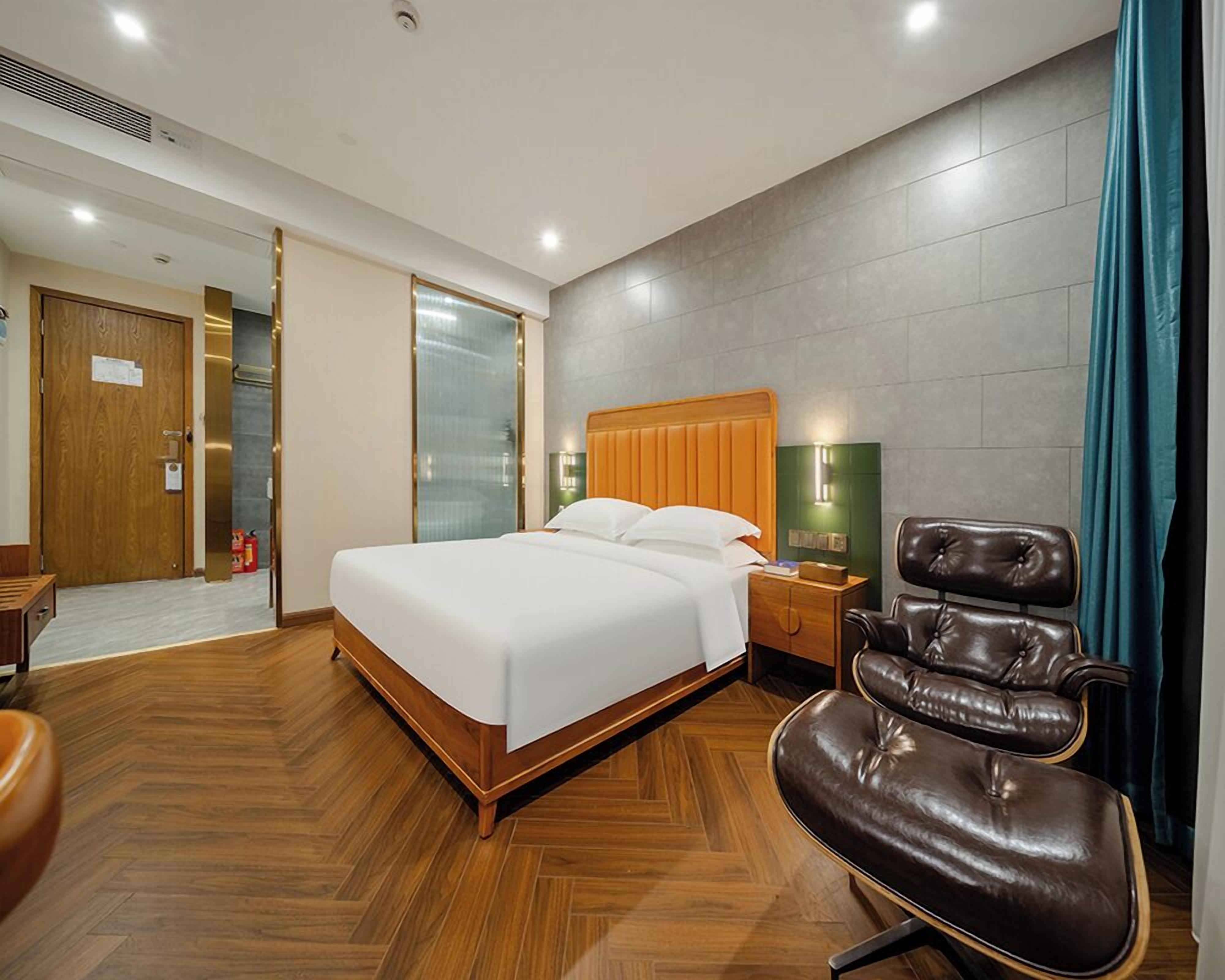 Ein Hotelzimmer mit Holzboden, einem Bett mit weißen Leinentüchern, einem Ledersessel, einem Nachttisch mit Lampe und einer grünen Wand mit Fenster.