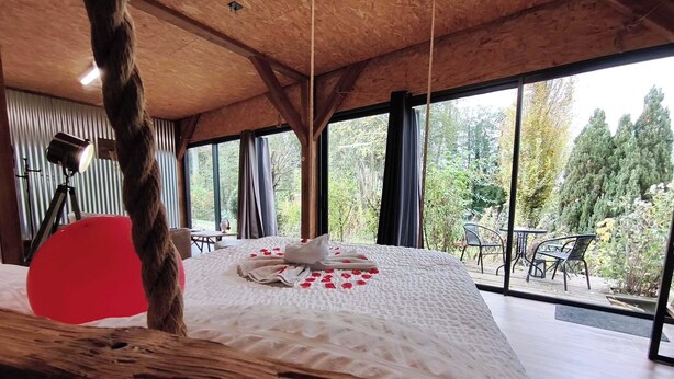 A bedroom with a large bed, a wooden ceiling, and a view of a garden through large windows. - Le lodge du bassin, Au Calme Absolu (Lindry)