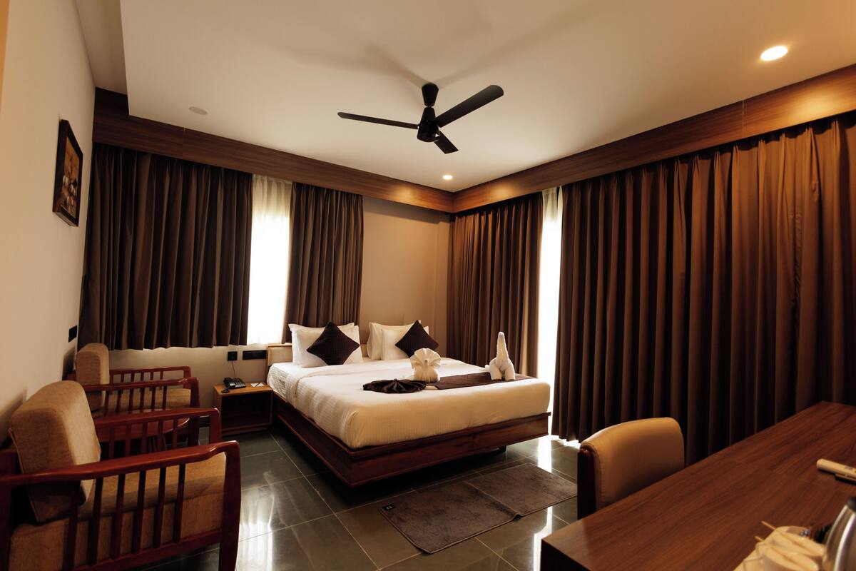 A hotel room with a large bed, two armchairs, a wooden desk, a ceiling fan, and a nightstand with a lamp.