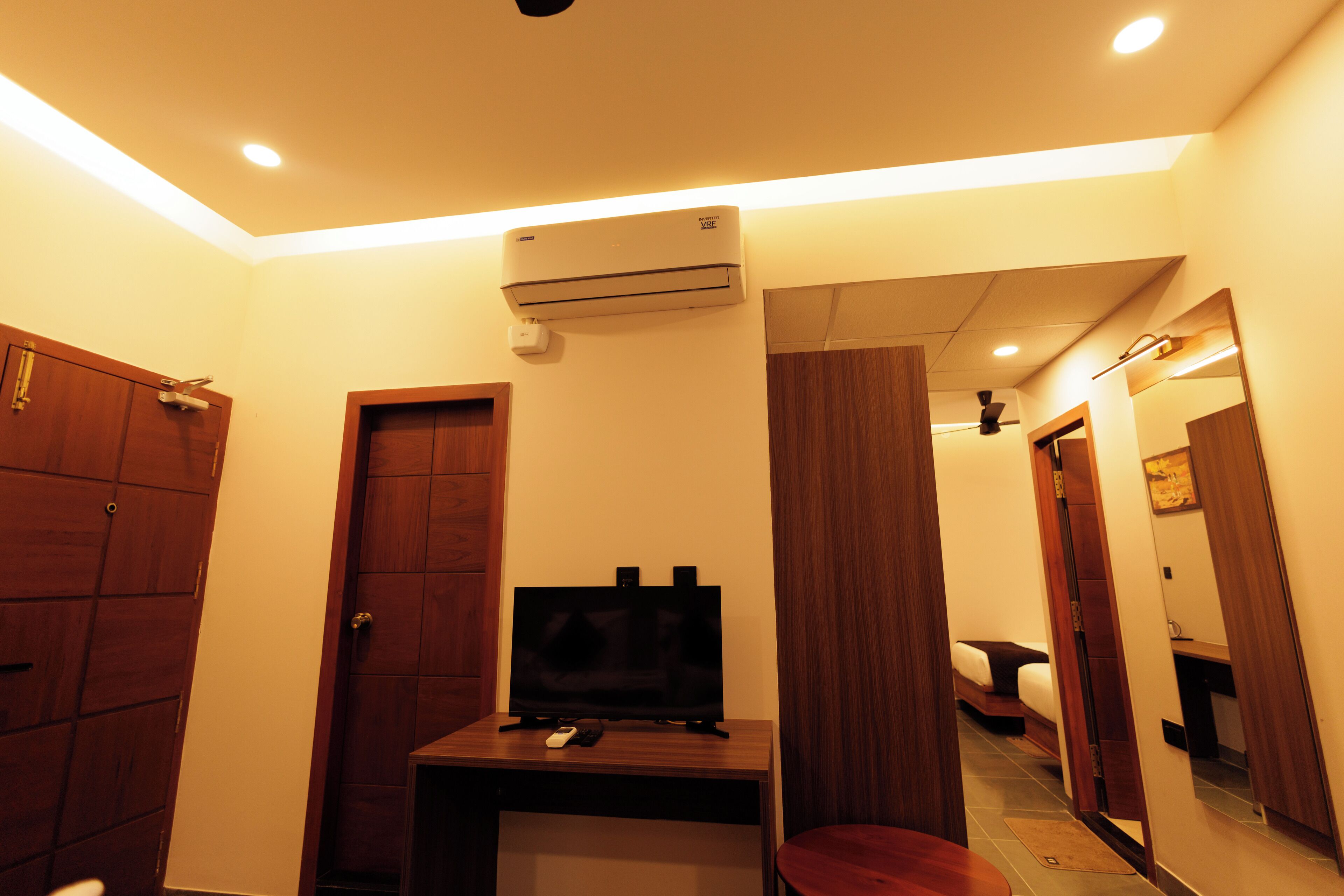 A hotel room with a wooden door, a flat-screen TV on a wooden stand, an air conditioning unit, and a mirror reflecting the room.