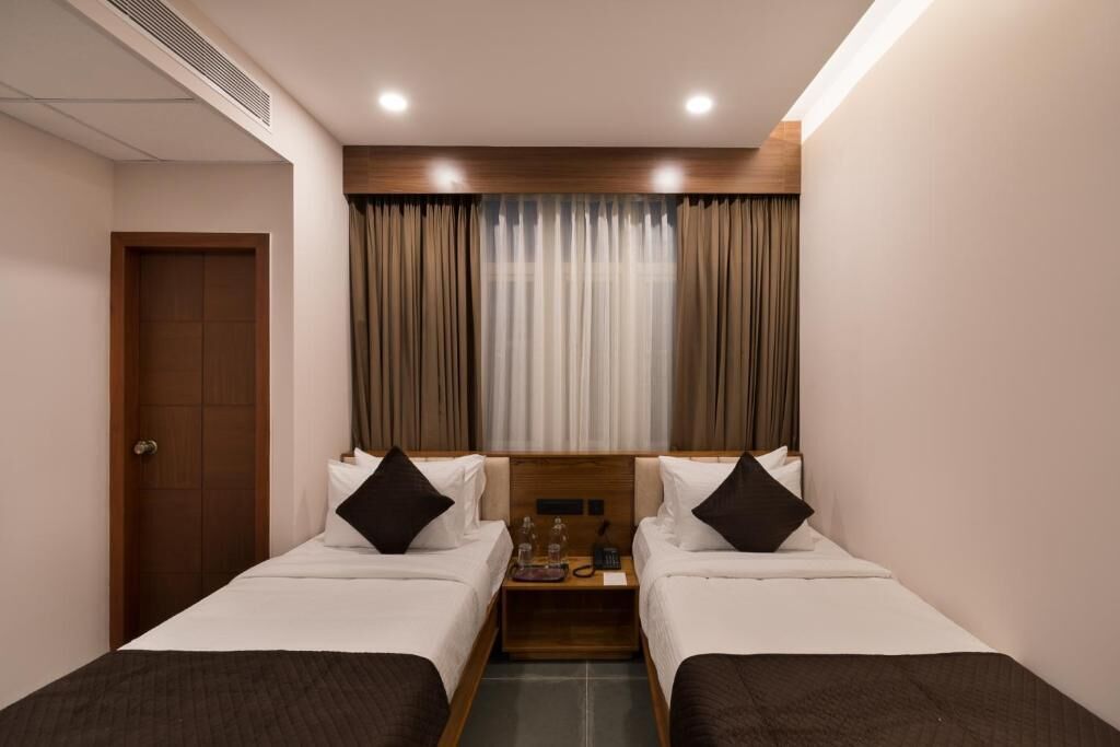 Quarto de hotel com duas camas, cabeceira de madeira, mesa de cabeceira com abajur e janela com cortinas.