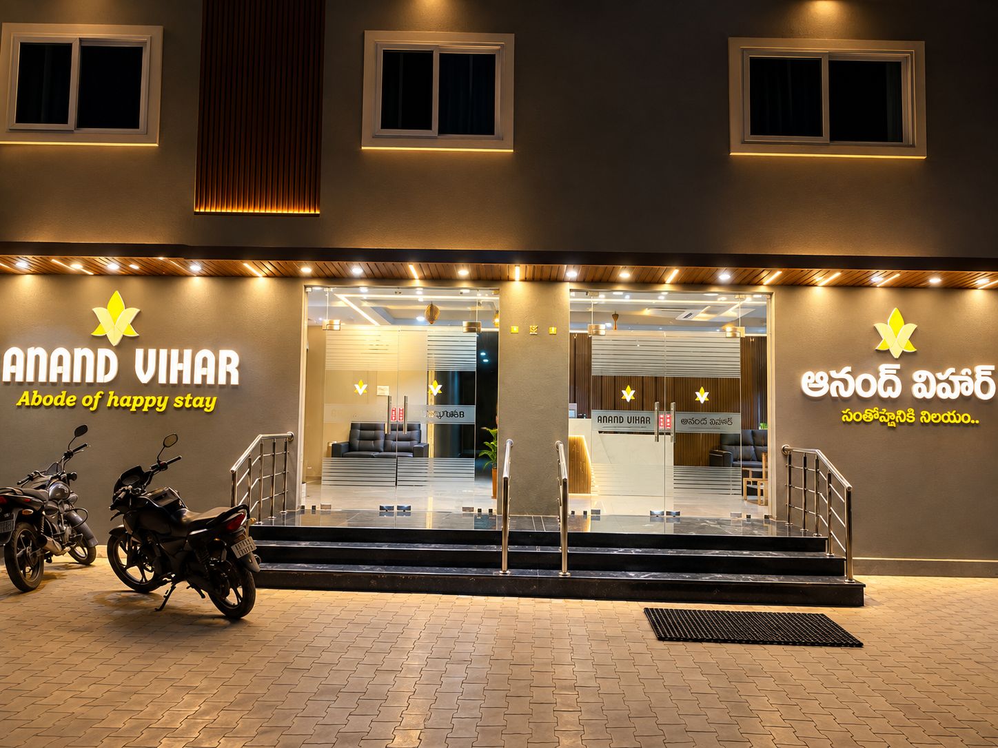 A hotel entrance with a sign reading 'Anand Vihar' and a logo. There are steps leading to the entrance, a motorcycle parked outside, and a wall-mounted sign with text in a local language.