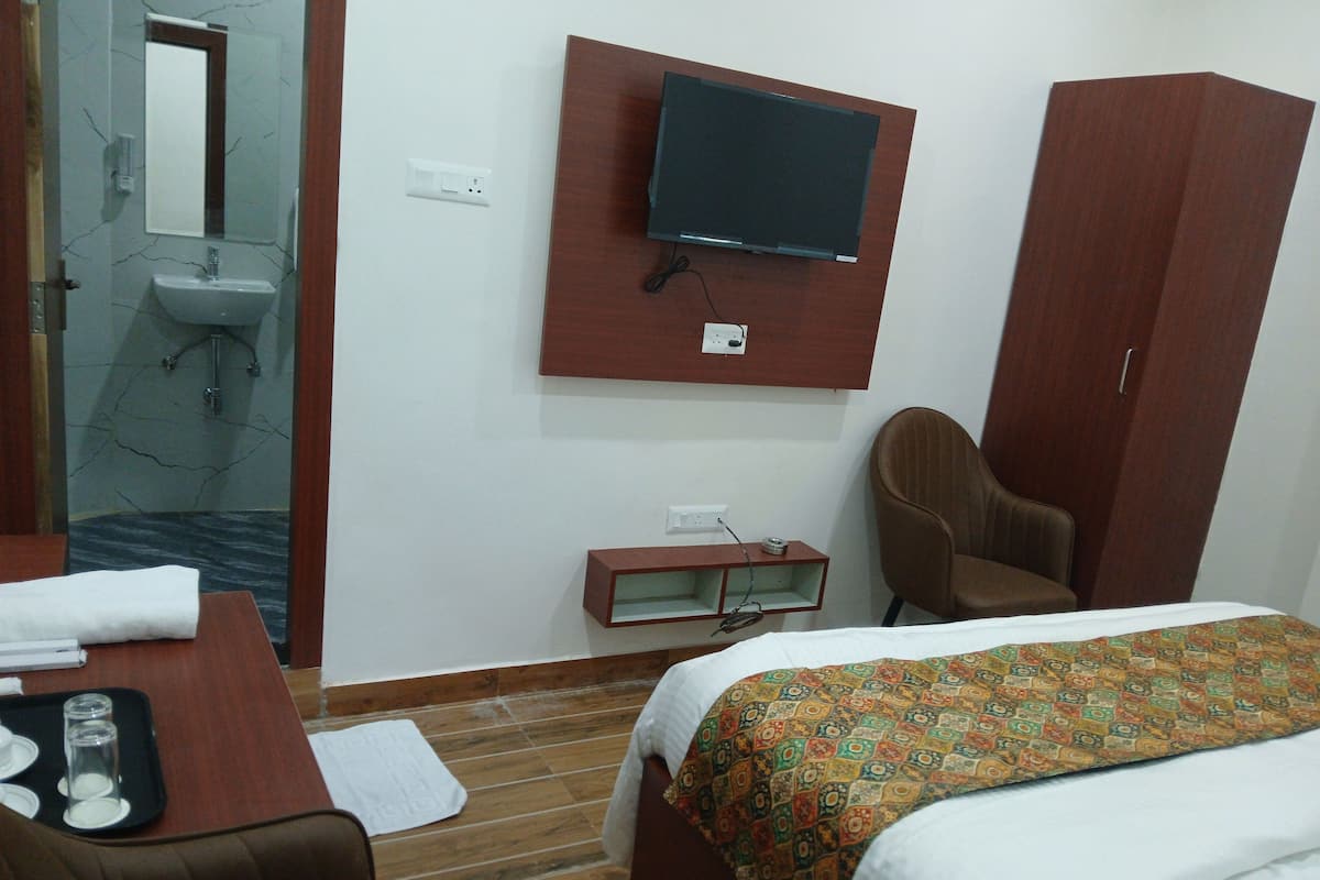 A hotel room with a bed, a desk with a chair, a television mounted on the wall, a wardrobe, and a bathroom with a sink and mirror.