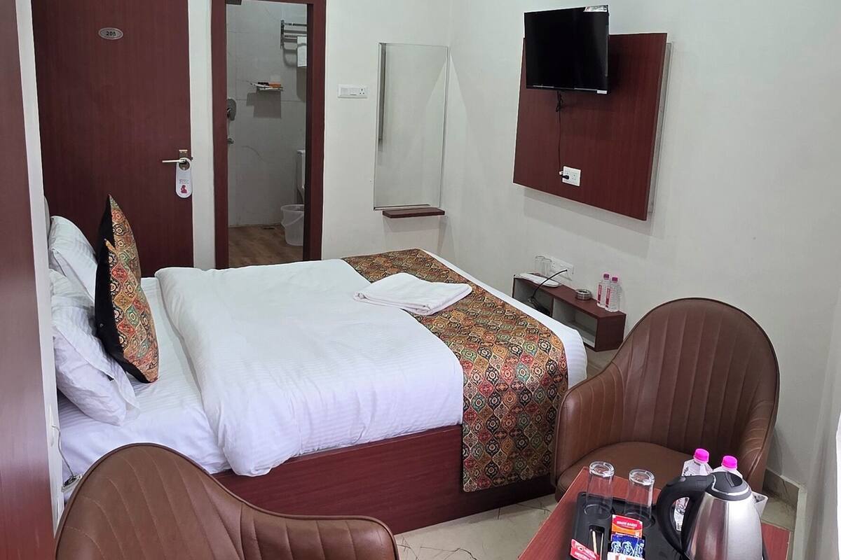 A hotel room with a bed, a desk with a kettle, a chair, a TV, and an air conditioner.