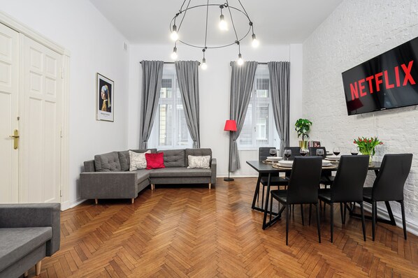 Una sala moderna con un sofá gris, una mesa de comedor con sillas negras y un televisor de pantalla plana mostrando "NETFLIX".