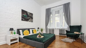 A modern bedroom with a herringbone wooden floor, a grey bed with a green blanket, a white nightstand with a lamp, and a green armchair. - Lumina bright apart near the city center (Łódź)
