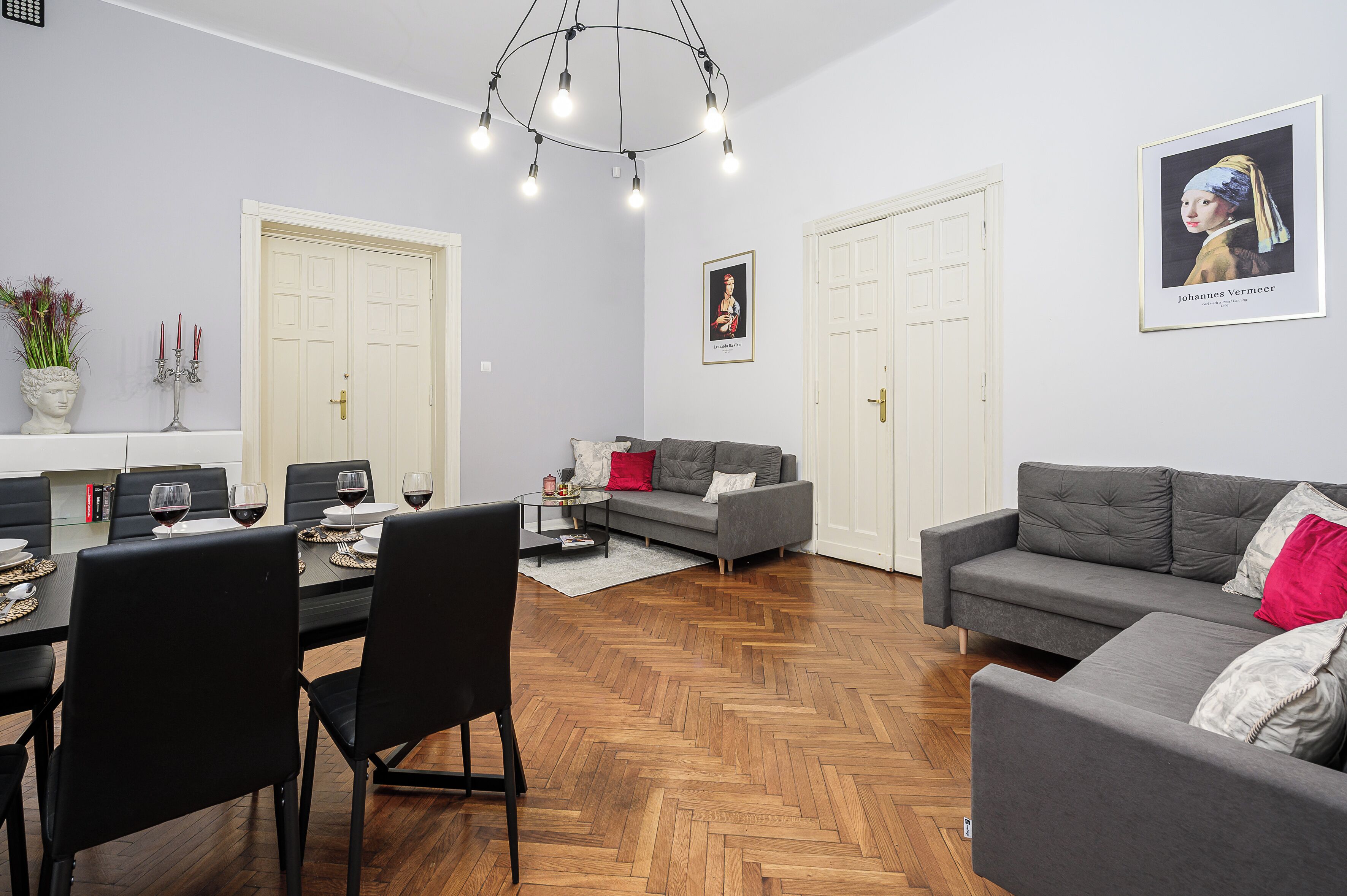 A modern living room with a dining area, grey sofas, a chandelier, and a framed artwork on the wall.