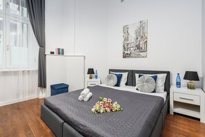 Una habitación de hotel moderna con una cama grande, una ventana con cortinas, una mesita de noche con lámpara, una mesita auxiliar y una obra de arte en la pared.