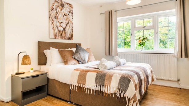 Ein Schlafzimmer mit Bett, Nachttisch, Lampe, Gemälde und Fenster mit Blick auf Grünfläche.