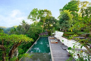 A pool with a wooden deck and lounge chairs, surrounded by lush greenery and trees. - Le Sunset by Nakula (Ubud)