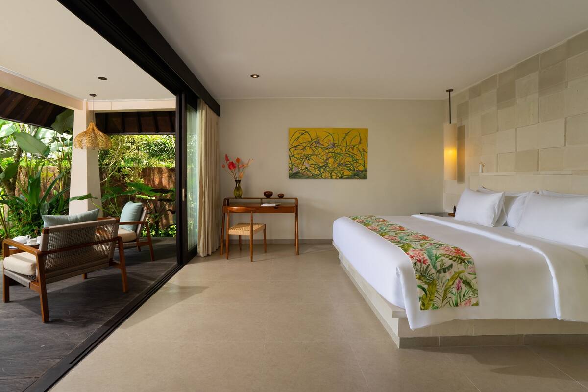 A modern bedroom with a large bed, a desk, chairs, and a view of greenery.